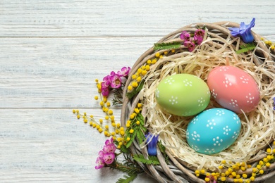 Wicker nest with painted Easter eggs and flowers on wooden table, top view. Space for text