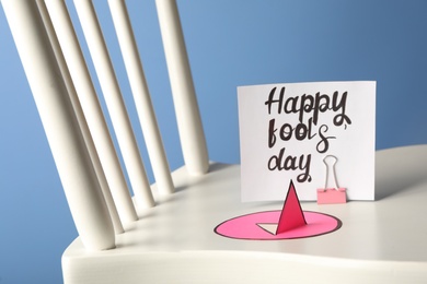 Photo of Chair with paper pin and Happy Fools' Day note on blue background, closeup