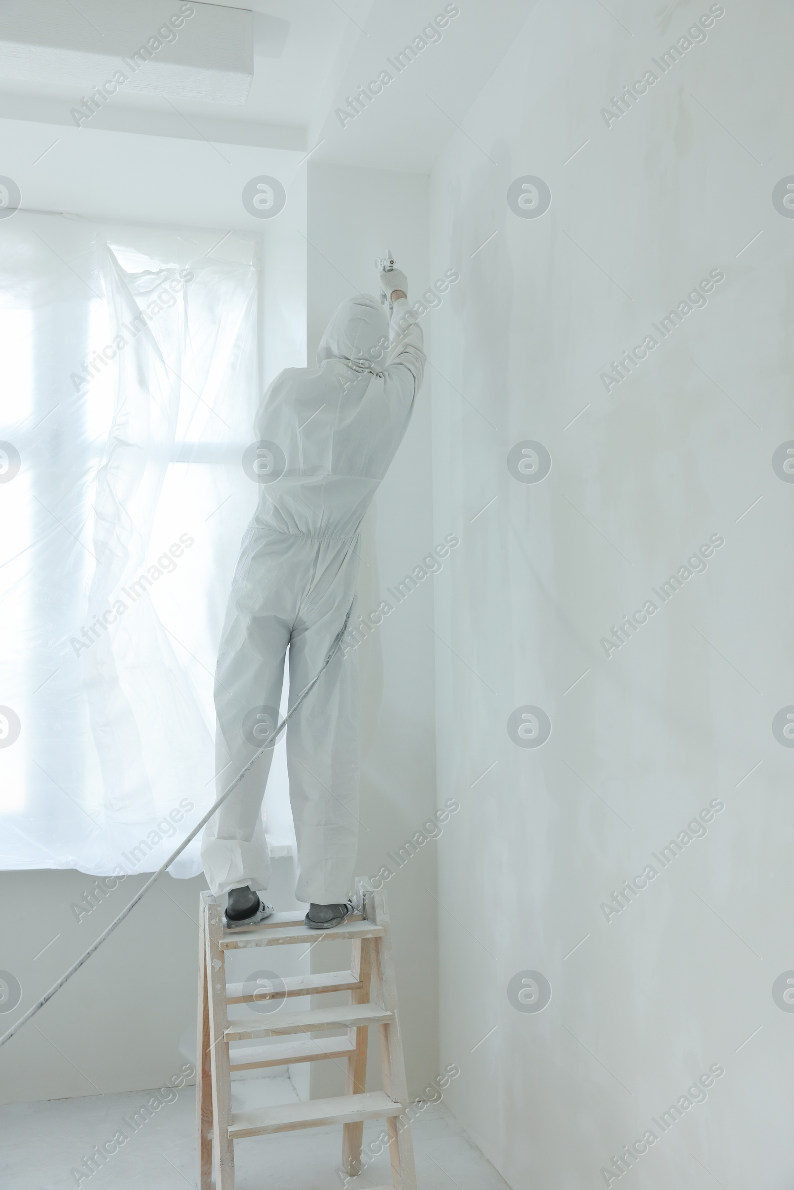 Photo of Decorator painting wall on ladder near windows indoors, back view