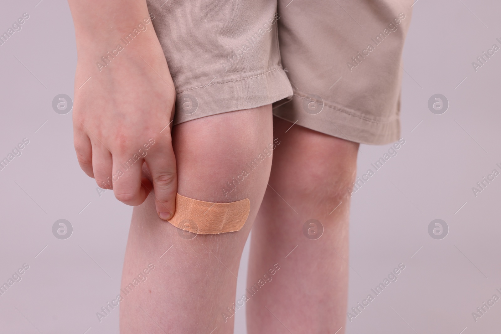 Photo of Little boy putting sticking plaster onto knee against light grey background, closeup