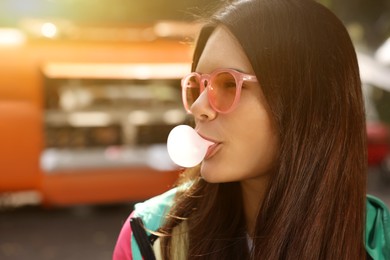 Photo of Beautiful young woman in pink glasses blowing chewing gum outdoors, space for text