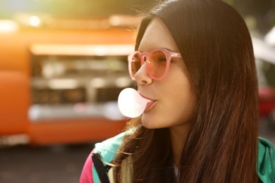 Beautiful young woman in pink glasses blowing chewing gum outdoors, space for text