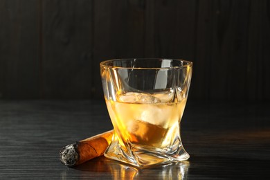 Glass of whiskey with ice cubes and smoldering cigar on black wooden table, closeup