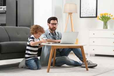 Photo of Man working remotely at home. Father using laptop while his son drawing at desk