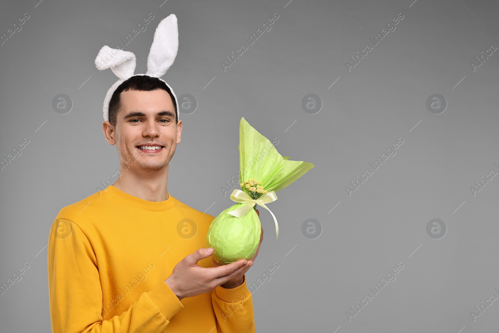 Photo of Easter celebration. Handsome young man with bunny ears holding wrapped gift on grey background. Space for text