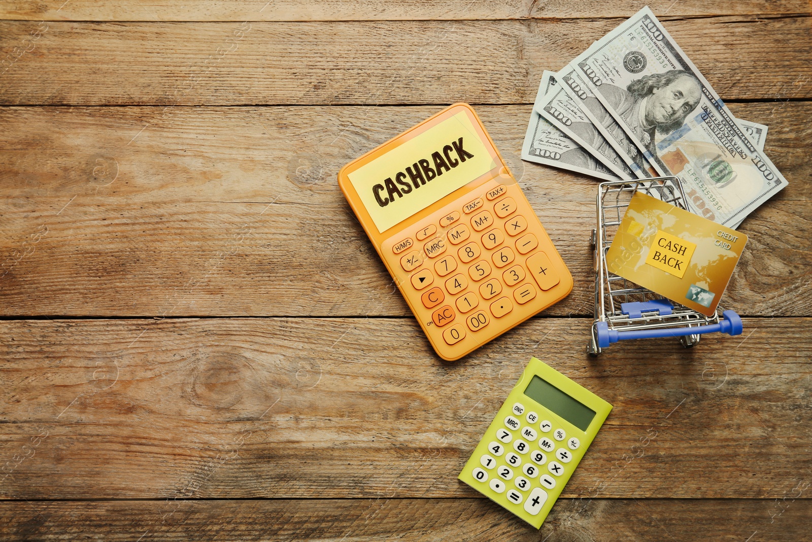 Photo of Calculators, dollar banknotes and credit card in shopping cart on wooden background, flat lay with space for text. Cashback concept