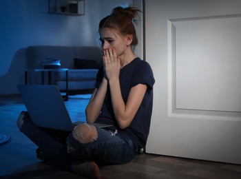 Photo of Terrified teenage girl with laptop on floor in dark room. Danger of internet