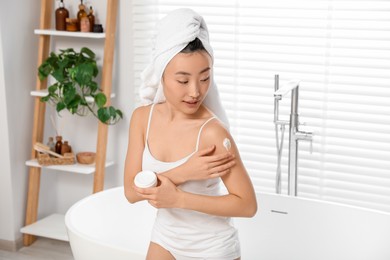 Photo of Beautiful young Asian woman applying body cream on shoulder in bathroom