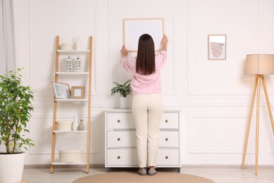 Woman hanging picture frame on white wall at home, back view