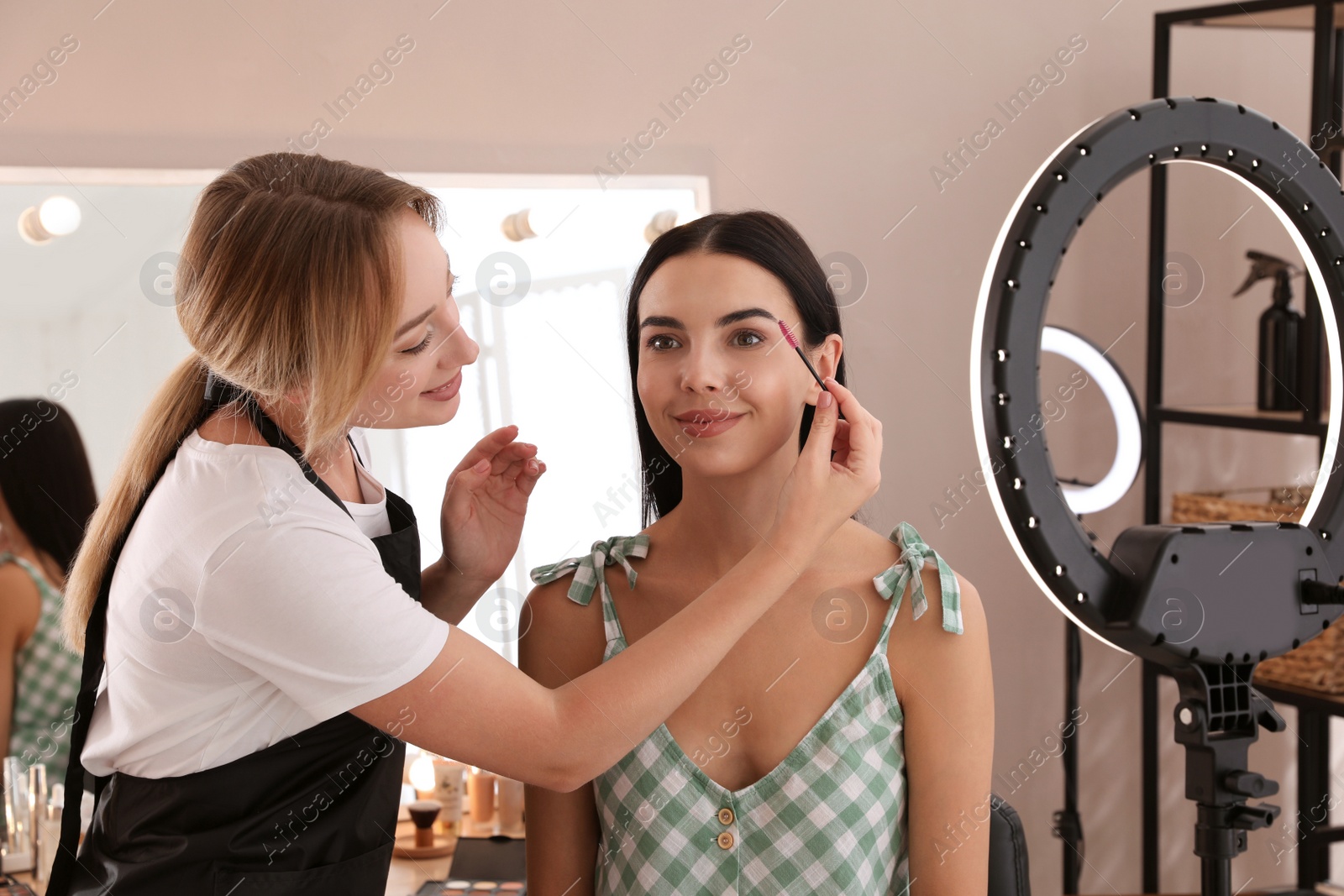 Photo of Professional makeup artist working with beautiful young woman in salon. Using ring lamp