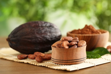 Photo of Wooden bowls of cocoa beans and powder near pod on table against blurred green background