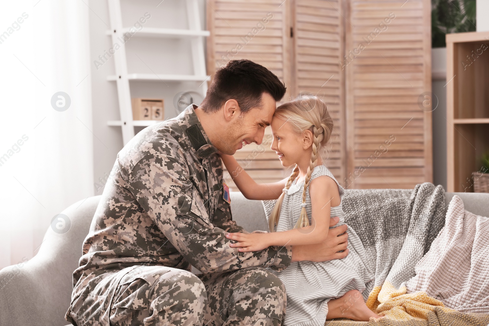 Photo of Young man in military uniform with his little daughter on sofa at home