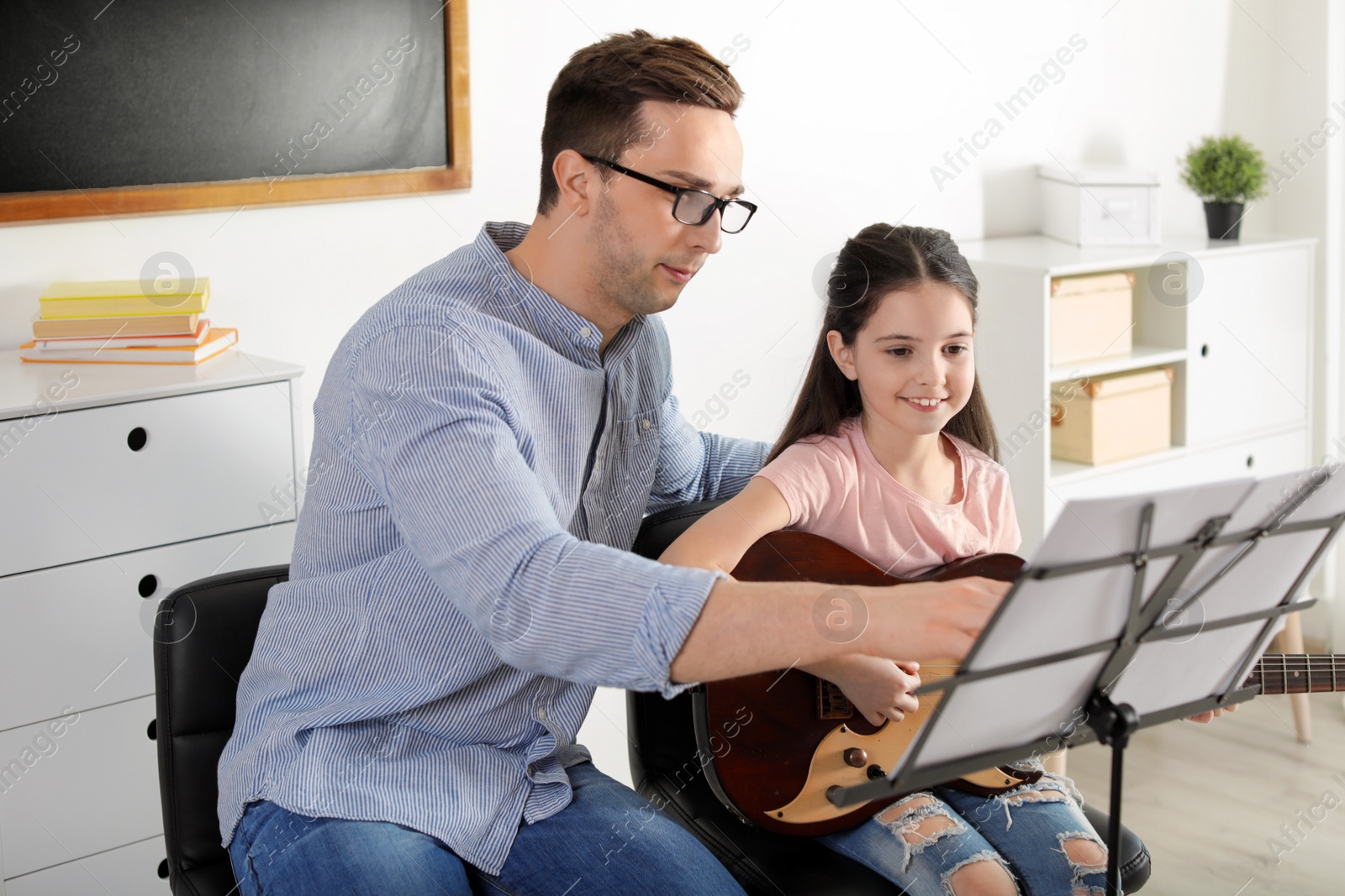 Photo of Little girl playing guitar with her teacher at music lesson. Learning notes