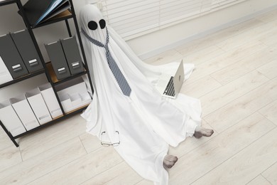 Photo of Overworked ghost. Man in white sheet with laptop on floor in office, above view