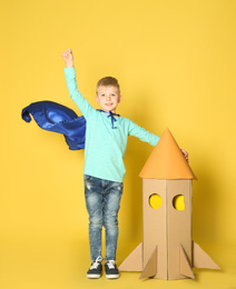 Little child in cape playing with rocket made of cardboard box on yellow background