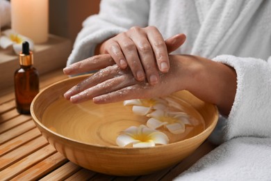 Photo of Woman applying scrub onto her hands in spa, closeup. Bowl of water and flowers on wooden table