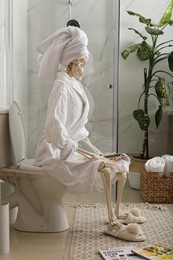 Skeleton in bathrobe with mobile phone sitting on toilet bowl