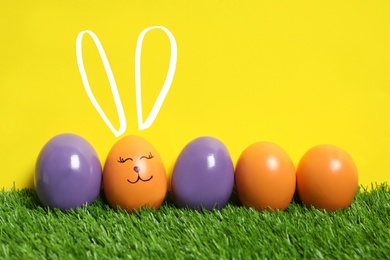 Image of One egg with drawn face and ears as Easter bunny among others on green grass against yellow background