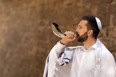 Photo of Jewish man blowing shofar on Rosh Hashanah outdoors. Wearing tallit with words Blessed Are You, Lord Our God, King Of The Universe, Who Has Sanctified Us With His Commandments, And Commanded Us To Enwrap Ourselves In Tzitziton in Hebrew