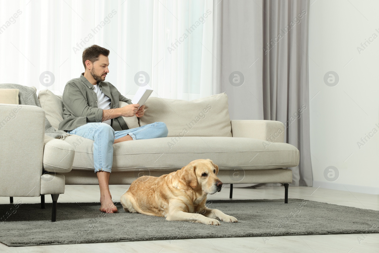 Photo of Man reading book on sofa near his cute Labrador Retriever at home. Space for text