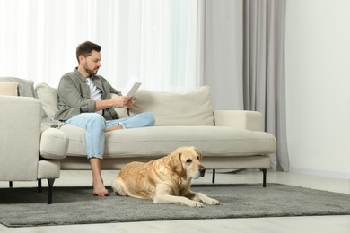 Photo of Man reading book on sofa near his cute Labrador Retriever at home. Space for text