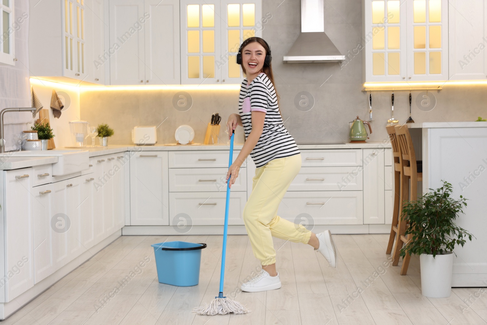 Photo of Enjoying cleaning. Happy woman in headphones dancing with mop in kitchen