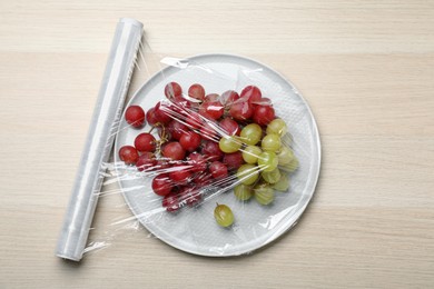 Photo of Plate of fresh grapes with plastic food wrap on wooden table, flat lay
