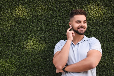 Young man with wireless headphones listening to music near green grass wall. Space for text