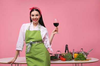 Photo of Young housewife with glass of wine, vegetables and different utensils on pink background. Space for text