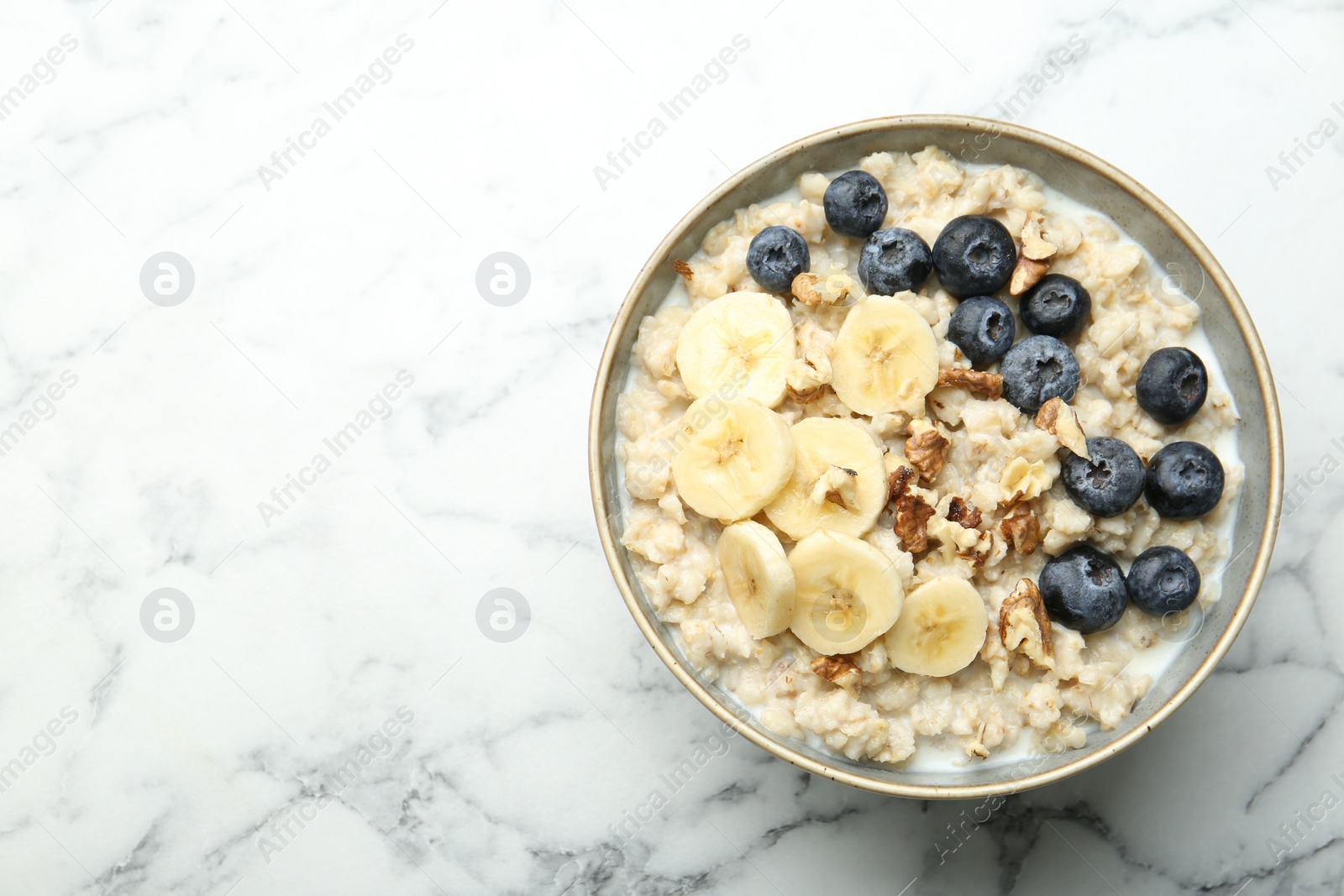 Photo of Tasty oatmeal with banana, blueberries, walnuts and milk served in bowl on white marble table, top view. Space for text