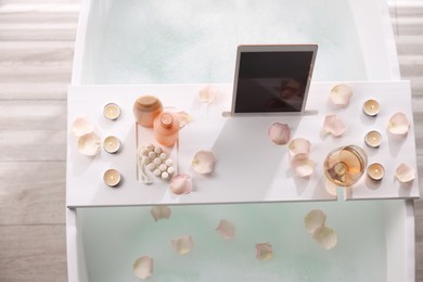Photo of Wooden tray with tablet, wine, toiletries and flower petals on bathtub in bathroom, above view