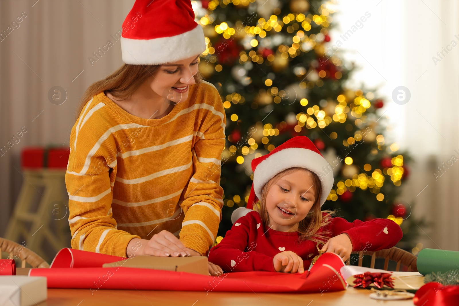 Photo of Mother and her little daughter in Santa hats wrapping Christmas gift at home