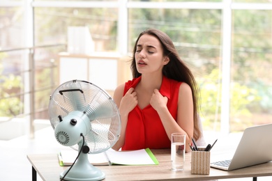 Photo of Young woman with fan suffering from heat at workplace. Summer season