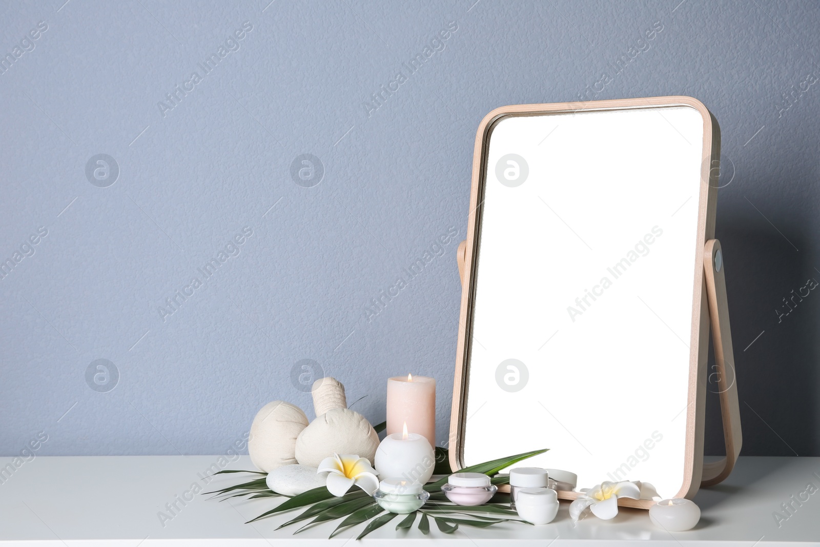 Photo of Stylish mirror, cosmetic products and aroma candles on table near light wall