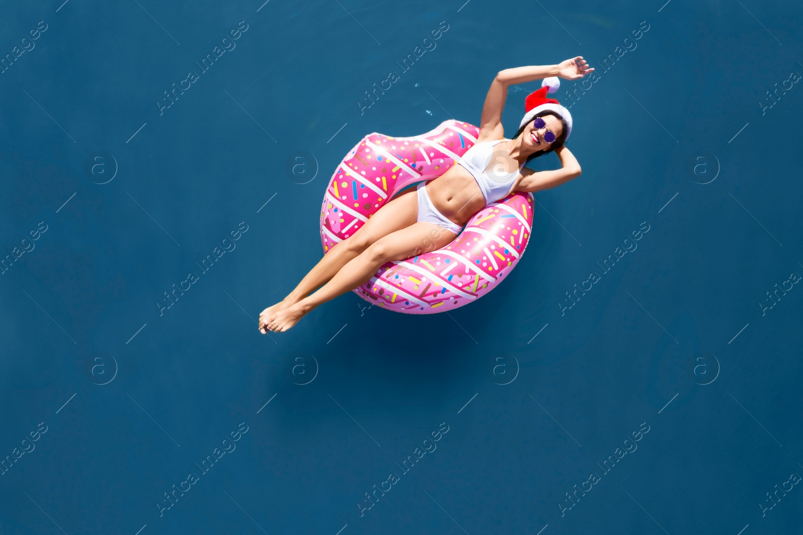 Image of Young woman wearing Santa hat and bikini with inflatable ring in sea, top view. Christmas vacation