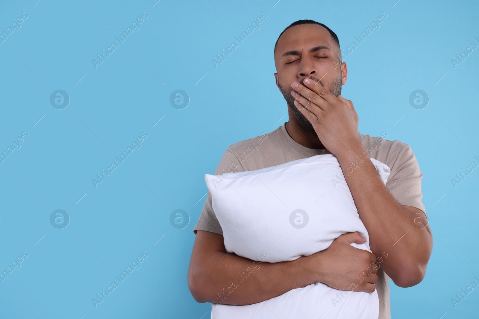 Photo of Tired man with pillow yawning on light blue background, space for text. Insomnia problem