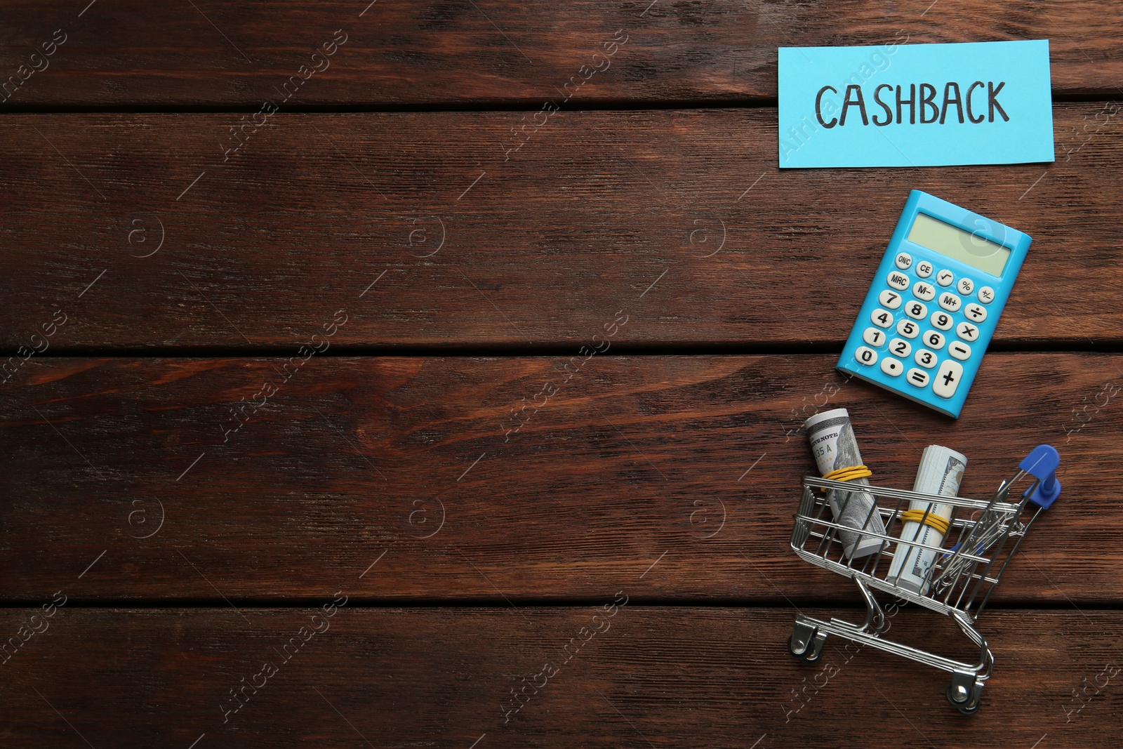 Photo of Card with word Cashback, calculator rolled dollar banknotes in shopping cart on wooden background, flat lay. Space for text