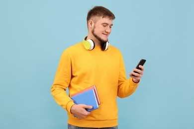 Young student with headphones and books using smartphone on light blue background