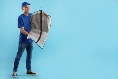 Dry-cleaning delivery. Happy courier holding garment cover with clothes on light blue background, space for text