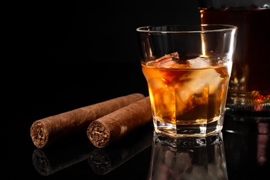 Photo of Glass of whiskey with ice cubes and cigars on black mirror surface, closeup