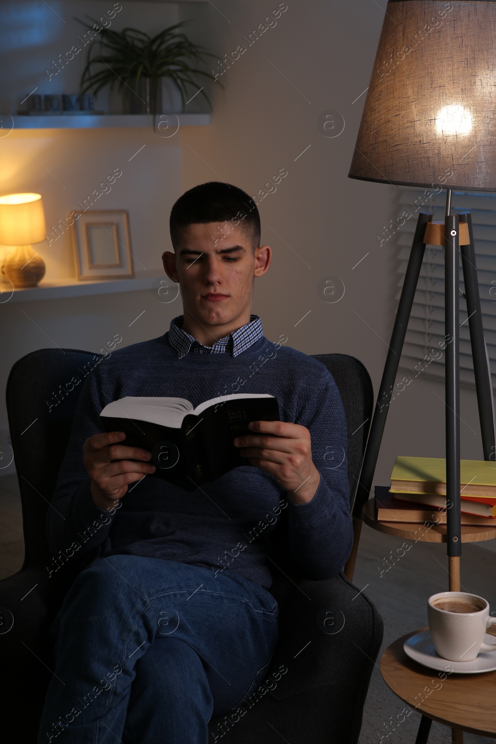 Photo of Young man with cup of drink reading book in cozy room at night