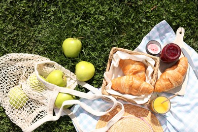 Photo of Blanket with juice, jam and croissants for picnic on green grass, flat lay
