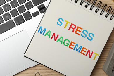 Image of Notepad with text STRESS MANAGEMENT and laptop on wooden table, flat lay