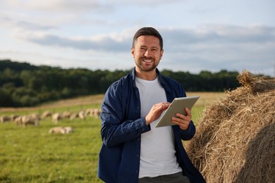 Photo of Smiling farmer with tablet near hay bale on pasture. Space for text