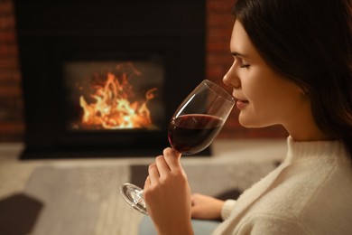 Young woman with glass of wine relaxing near fireplace at home. Space for text
