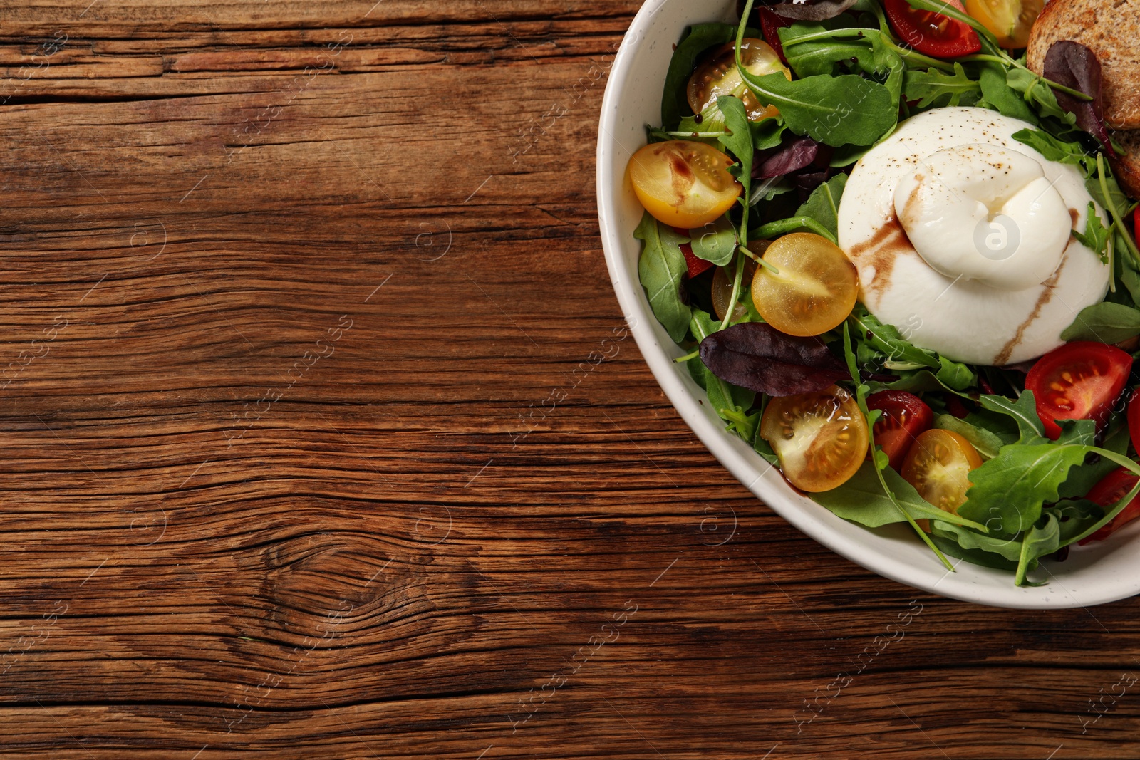 Photo of Delicious burrata salad with colorful cherry tomatoes, croutons and arugula on wooden table, top view. Space for text