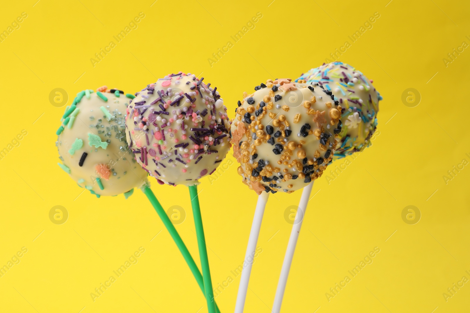 Photo of Sweet cake pops decorated with sprinkles on yellow background, closeup. Delicious confectionery