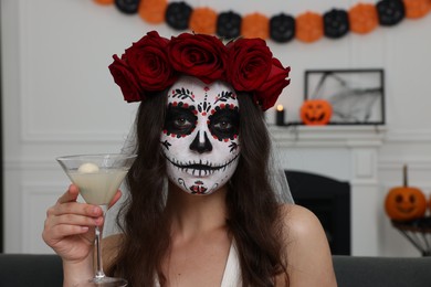 Young woman in scary bride costume with sugar skull makeup, flower crown and glass of cocktail indoors. Halloween celebration