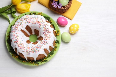 Photo of Glazed Easter cake with sprinkles, painted eggs and flowers on white wooden table, flat lay. Space for text