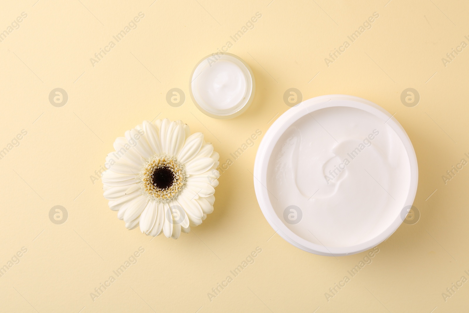 Photo of Moisturizing cream in open jars and gerbera flower on beige background, flat lay. Body care product